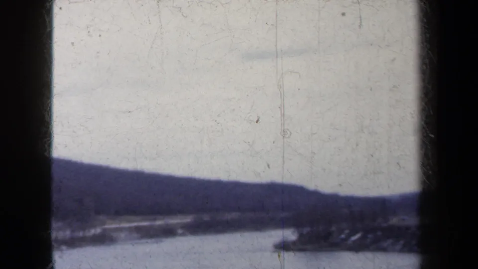 Faded 8mm winter landscape frame from New York, showing river water, a snow-dusted shoreline, and dark hills.