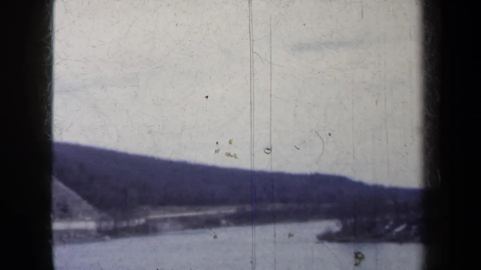1959 New York home-movie frame showing a lake, distant wooded ridge, and steep mountainside from a traveling car perspective.