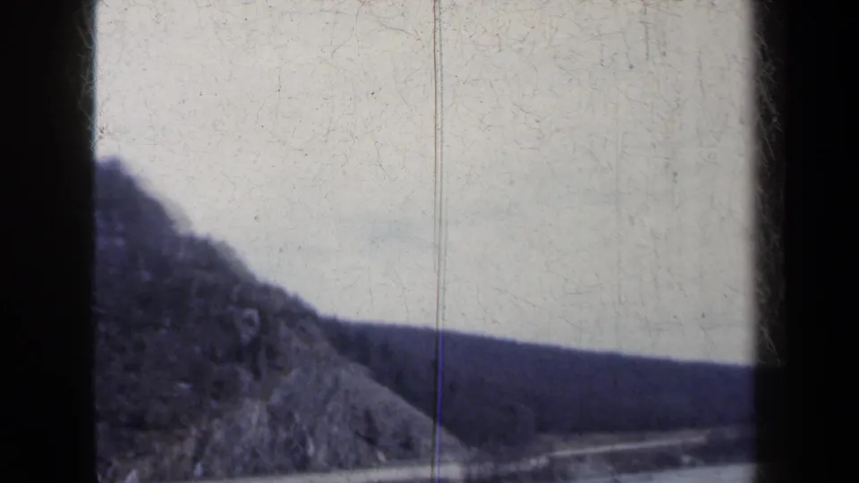 A steep forested hill rises beside a lake road in this 1959 New York 8mm travel shot.