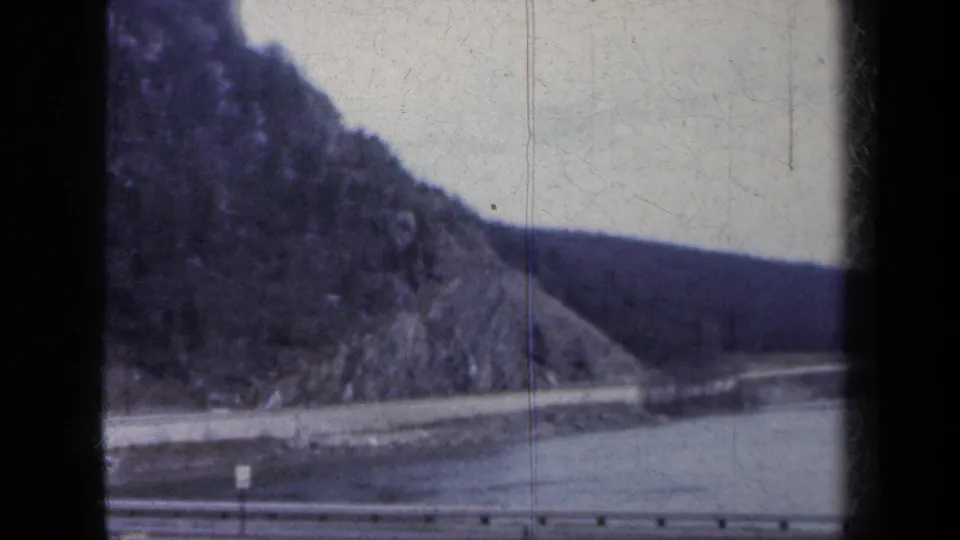 Moving 8mm frame of a guardrail, lakeside road, water, and rugged hill in New York, dated by metadata to 1959.