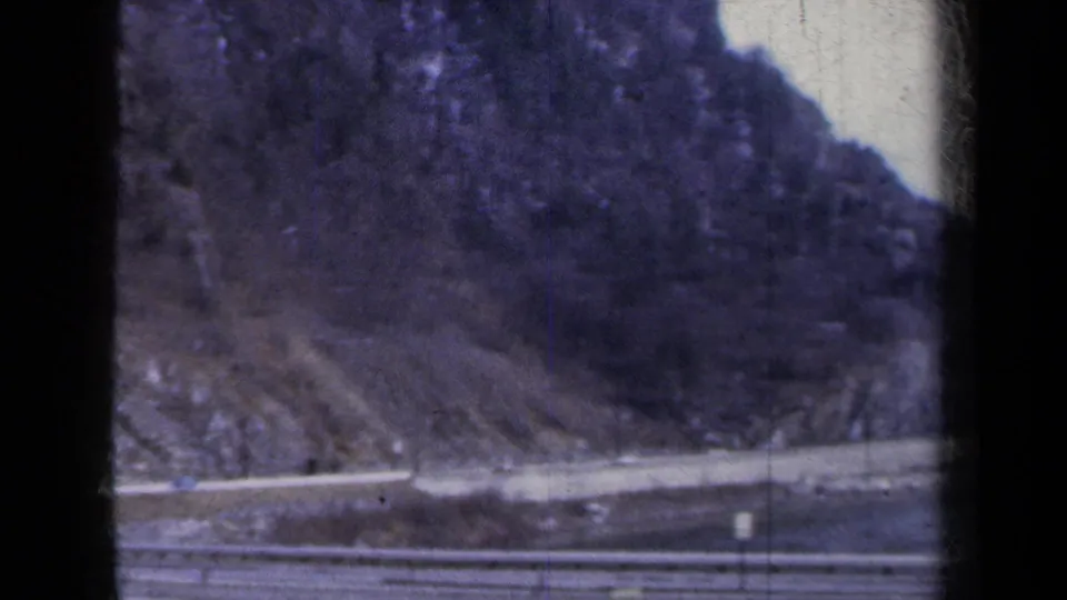 1959 New York home-movie view of a rocky lakeside hillside and road, framed by 8mm film borders and scratches.