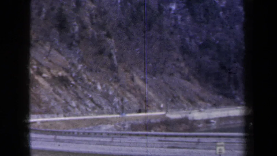 1959 New York home-movie frame of a winding highway ramp below rocky slopes and bare trees, partly obscured by dark foreground bars.
