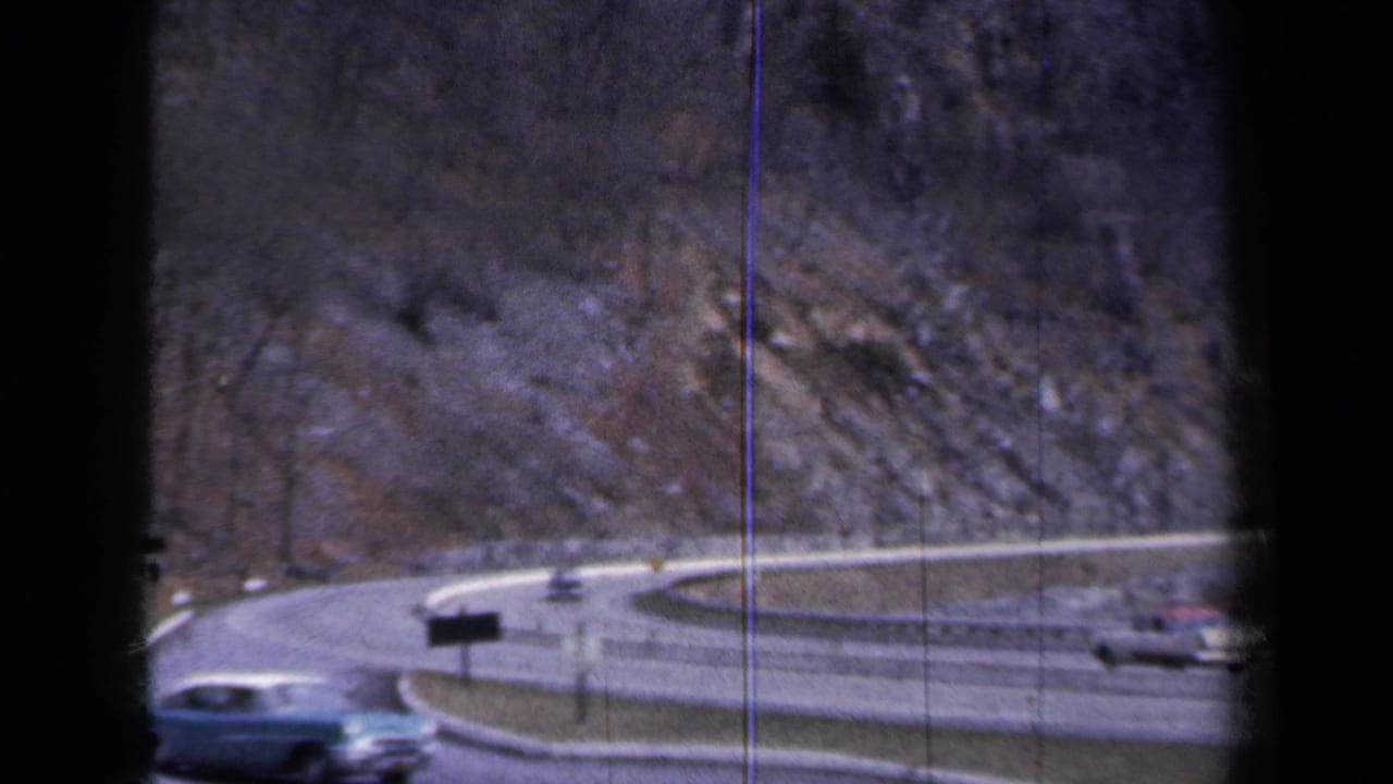 Light blue sedan passes a curved ramp beside rocky hillside and guardrails.