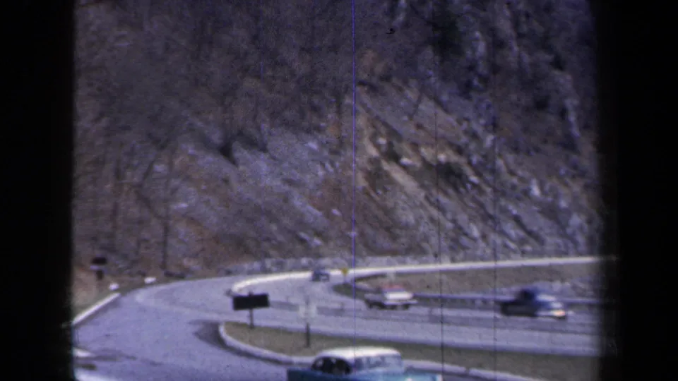 1959 New York home-movie view of several cars navigating a curved highway ramp, with signs, guardrails, and a rocky hillside behind.