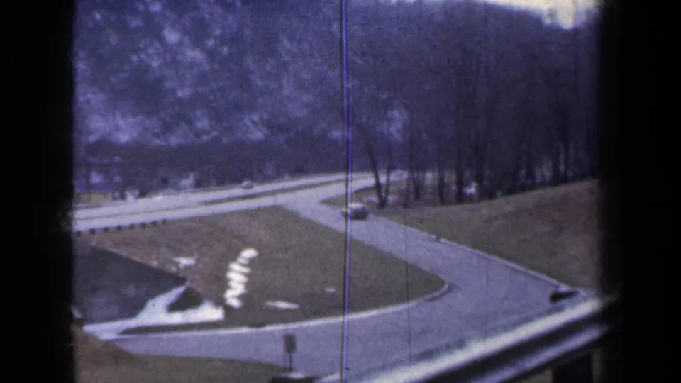 1959 New York frame showing elevated highway ramps, a grassy median, bare trees, patches of snow or pale ground, and a car at the edge.