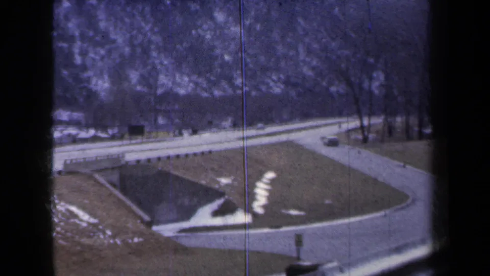 1959 New York 8mm frame showing a winter roadway, bridge structure, guardrails, bare trees, and snow patches from a moving vehicle.