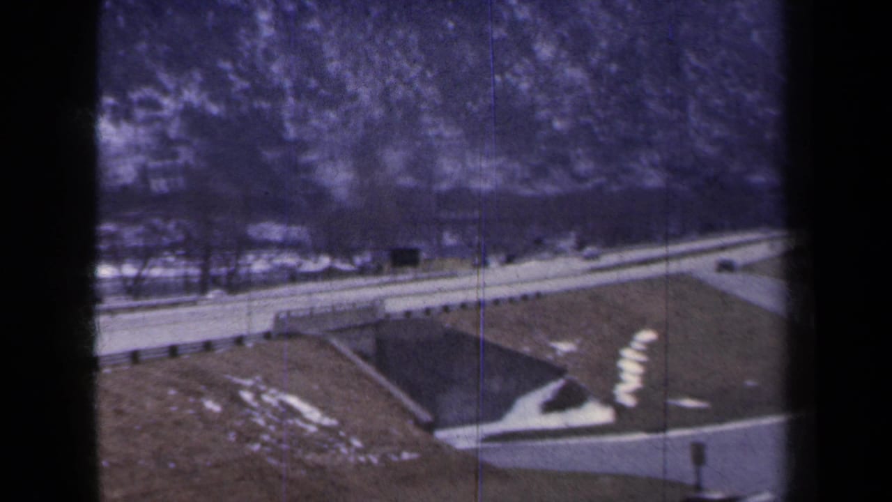 1959 New York home-movie frame with a road crossing near a bridge, winter snow patches, guardrails, and distant cars.