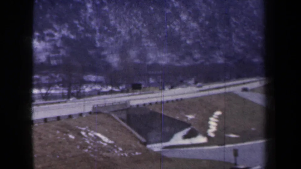 1959 New York home-movie frame with a road crossing near a bridge, winter snow patches, guardrails, and distant cars.