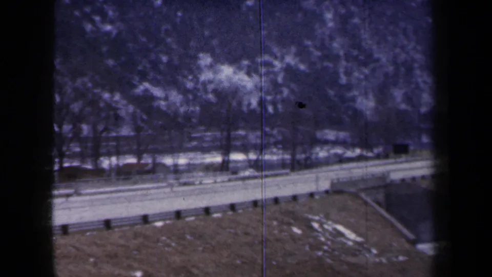 1959 New York 8mm frame showing a winter highway edge, guardrail, roadside snow, bare trees, and a bridge support area.
