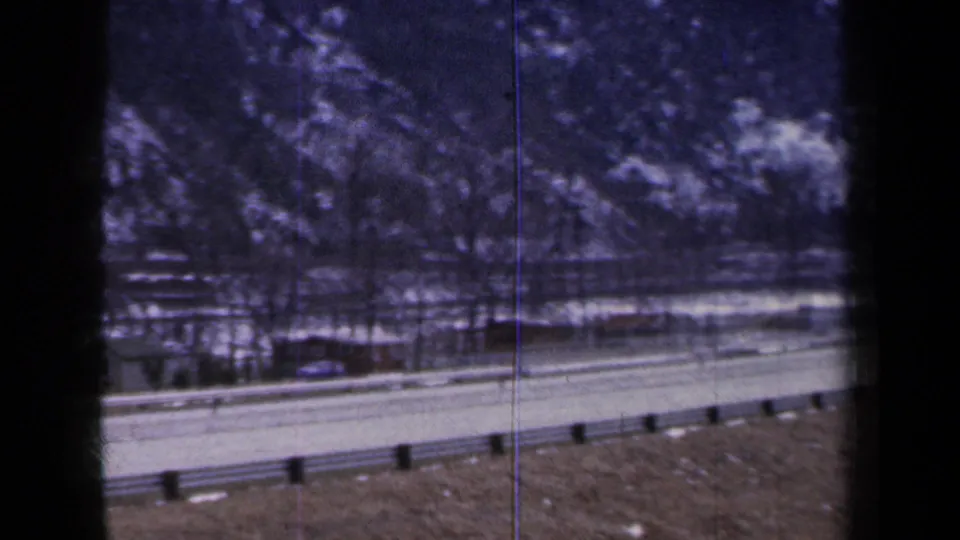 1959 New York home-movie frame of a winter road with guardrails, bare trees, snow patches, and a wooded slope beyond.
