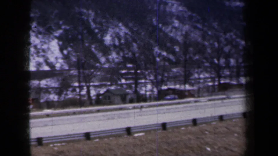 1959 New York 8mm frame showing a snowy highway shoulder, guardrail, bare trees, and distant roadside structures from a moving car.