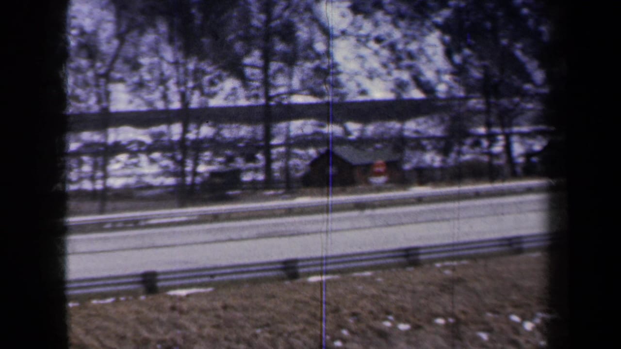 1959 New York 8mm view of a snowy roadside, guardrail, bare trees, and a small dark building seen from a moving vehicle.
