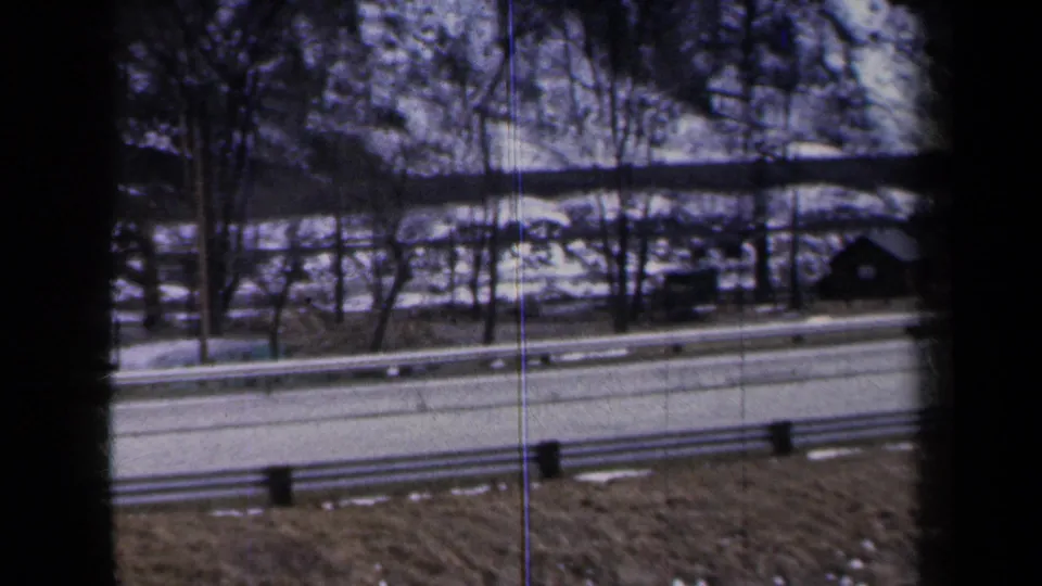 1959 New York home-movie frame looking across a guardrail toward snow, bare trees, and a small roadside building.