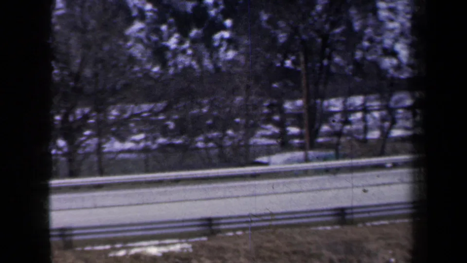 1959 New York 8mm frame of a snowy roadside and guardrail, softened by motion and framed by dark car-window or film edges.