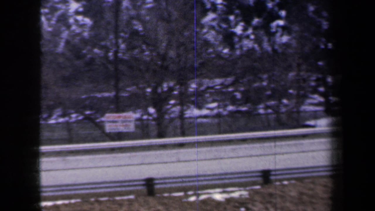 1959 New York 8mm frame showing a snowy highway shoulder, guardrails, trees, and a roadside sign from a moving car.