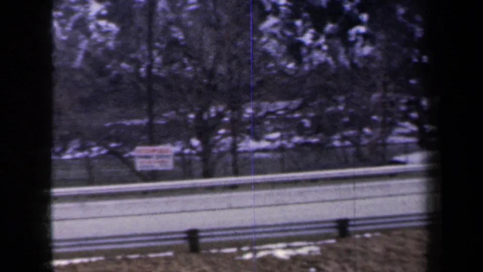 1959 New York 8mm frame showing a snowy highway shoulder, guardrails, trees, and a roadside sign from a moving car.