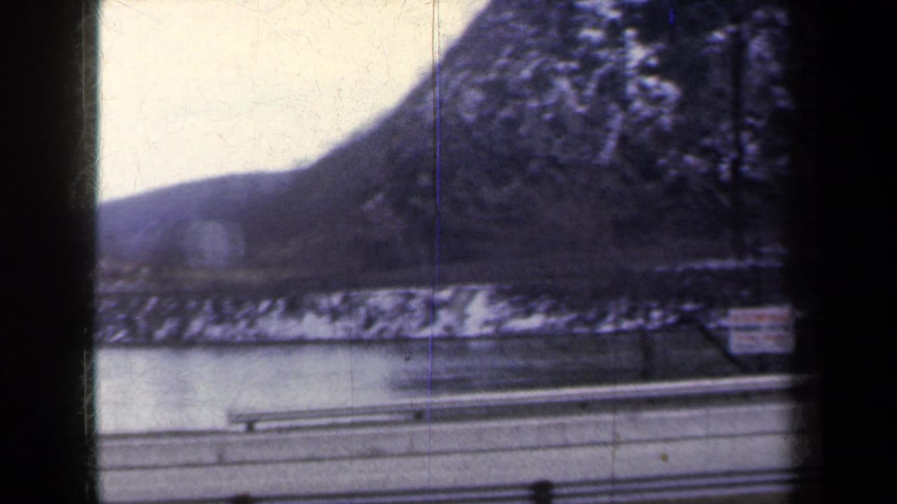 1959 New York 8mm frame with a guardrail, water, snow-dusted trees, and distant hills filmed from a moving vehicle.