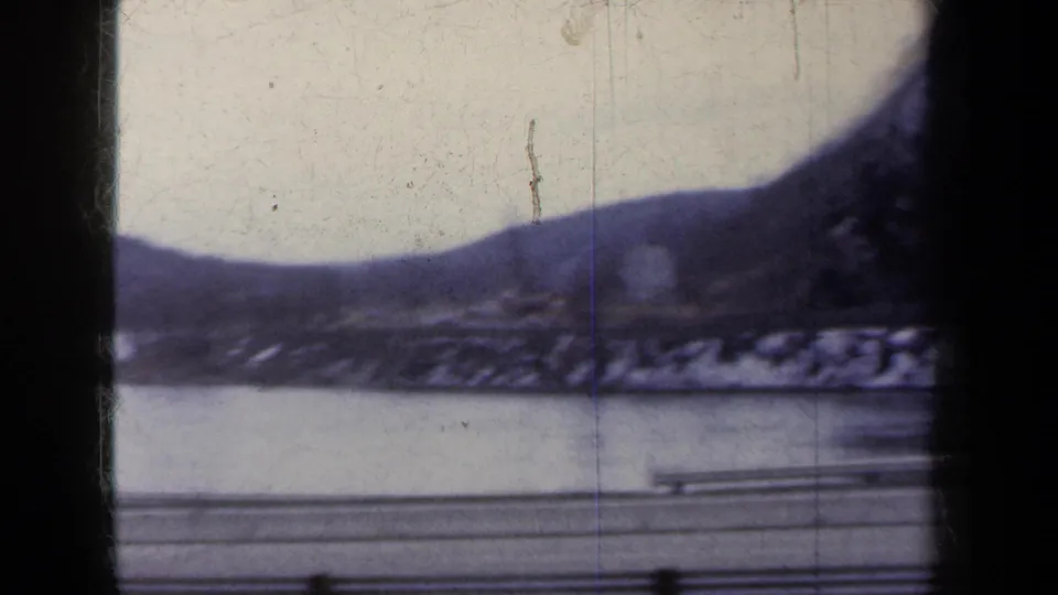 1959 New York 8mm frame looking past a winter road and guardrail toward water and low wooded hills.