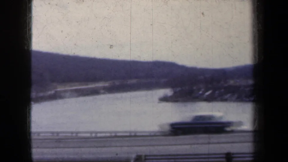 1959 New York 8mm frame showing a blurred car on a roadside near water, with snowy hills behind it.