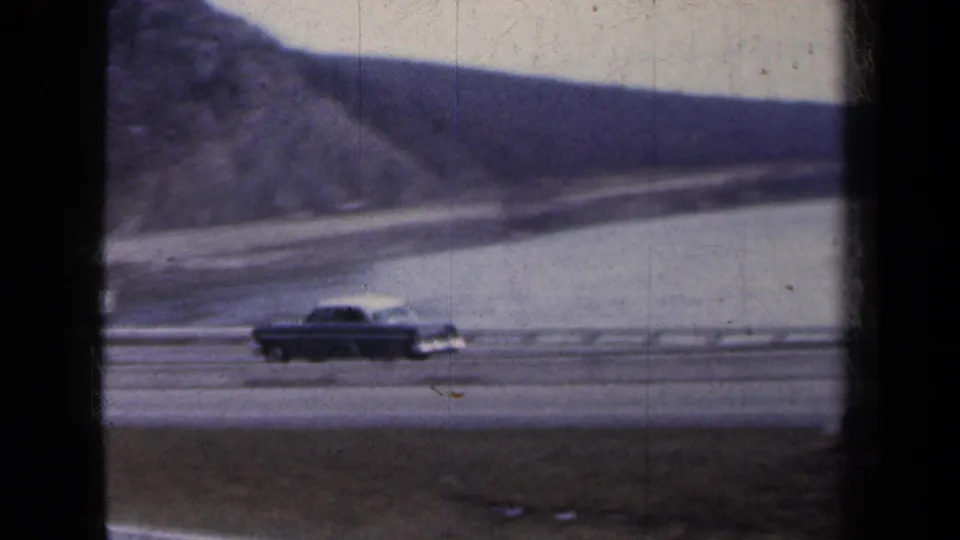 1959 New York 8mm frame showing a dark sedan on a highway beside calm water and distant hills, partly framed by a window edge.