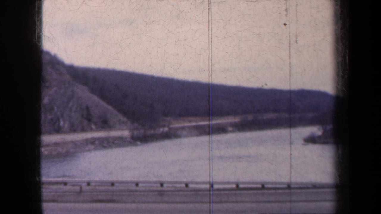 Passenger-view 8mm frame of a New York waterside highway, guardrail, calm water, and rugged hillside, with no people visible.