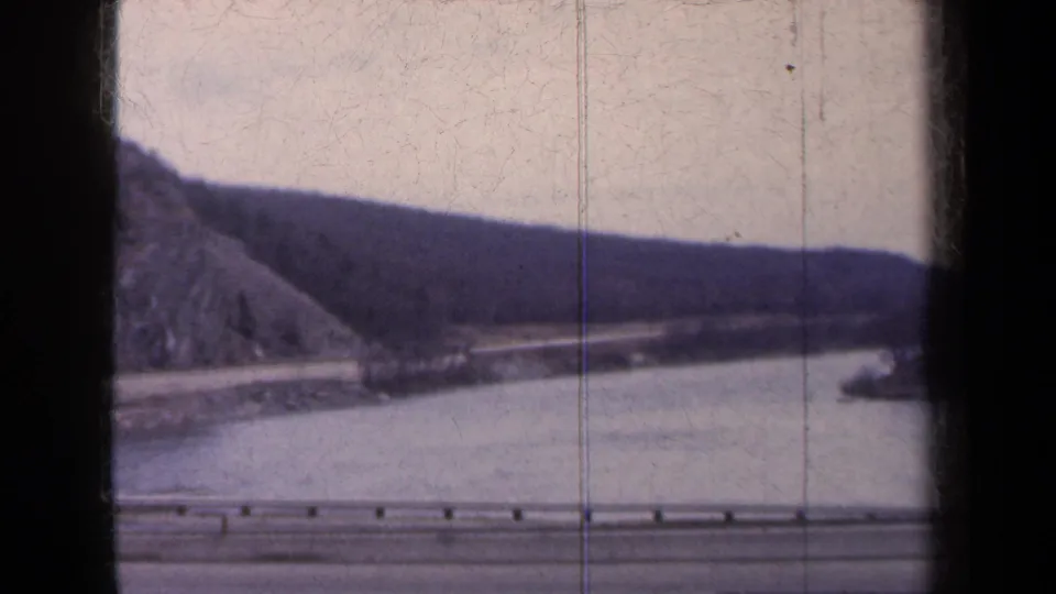 Passenger-view 8mm frame of a New York waterside highway, guardrail, calm water, and rugged hillside, with no people visible.