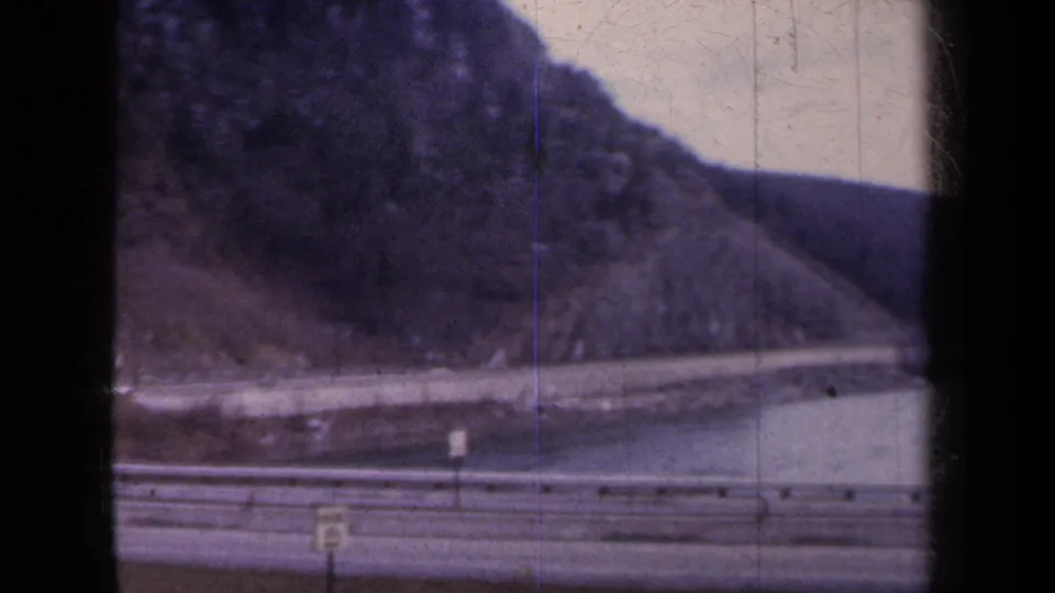 1959 home-movie frame showing a highway edge, guardrail, roadside sign, water, and steep hillside from a moving passenger viewpoint.