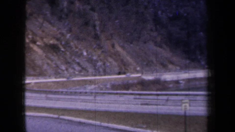 8mm frame from a 1959 New York road trip showing a highway barrier, small road signs, water, and rocky hillside through dark window edges.