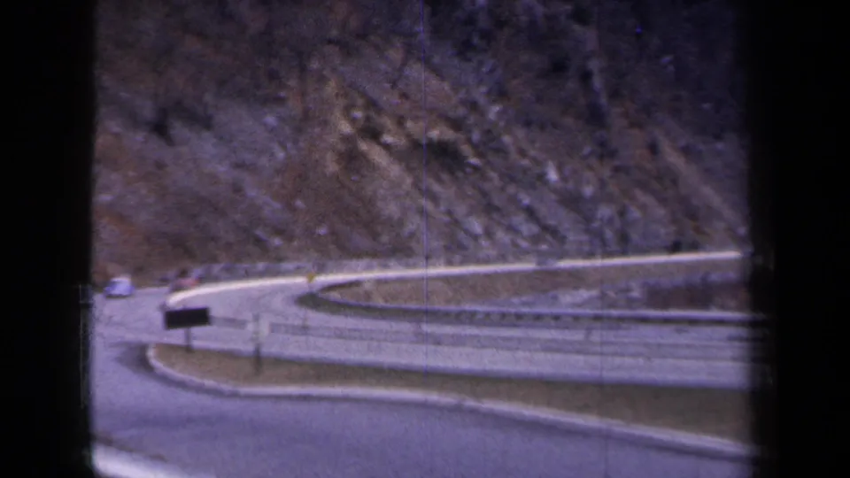 1959 New York home-movie frame showing a curving mountain road with guardrails, bare trees, rocky hillside, and a distant car.