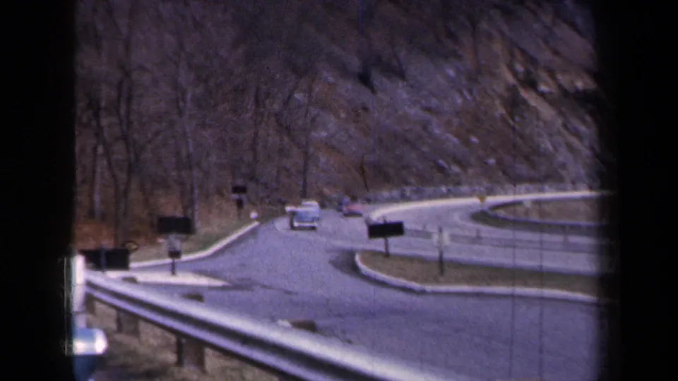 1959 New York road scene with classic cars moving through a guarded curve bordered by bare trees and rocky terrain.