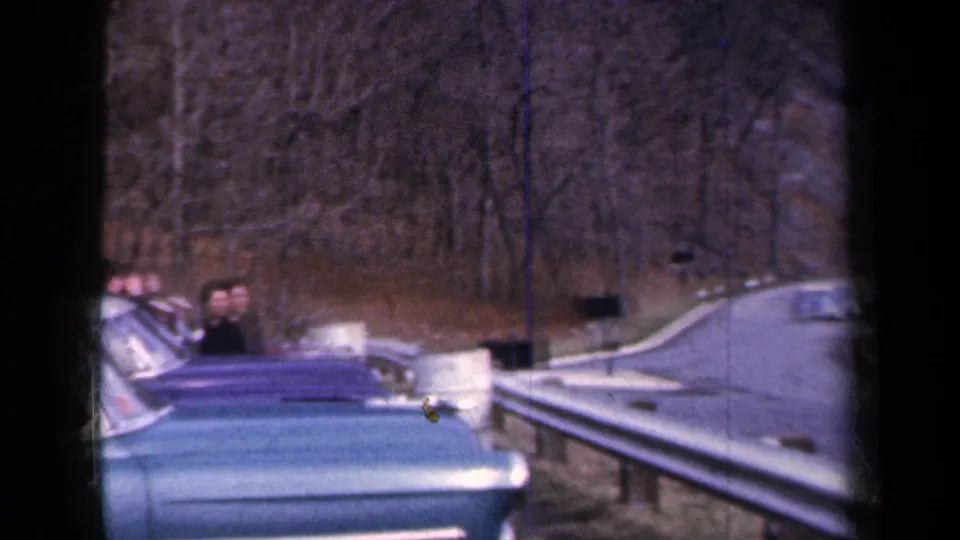 1959 New York home-movie frame with a blue convertible carrying visible passengers beside a curving guarded road.