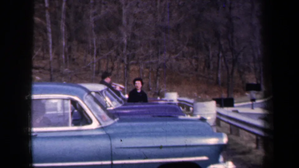 1959 New York 8mm frame showing blue mid-century cars stopped by a roadside guardrail, with two adults visible near the vehicles.