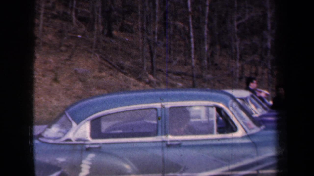 1959 New York roadside view of a blue mid-century car beside a metal guardrail, with wooded hillside in the background.