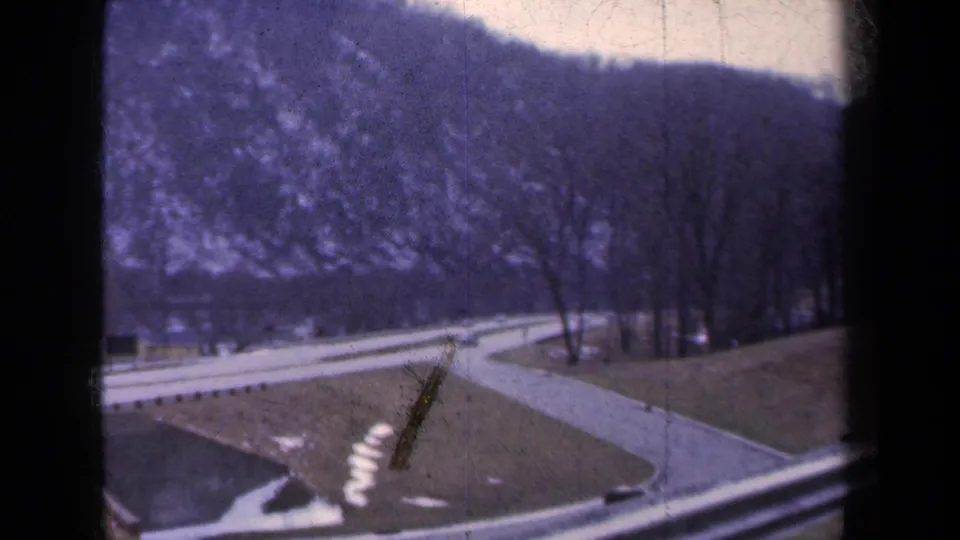 Moving 8mm view in New York showing a curving road, bare trees, hillside, and scattered snow patches in a roadside landscape.