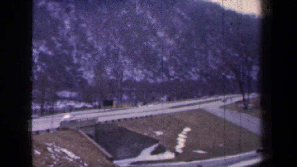 1959 New York home-movie frame of a roadside landscape with a curving road, bare trees, low hillside, and small snow patches.