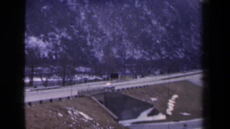 1959 New York 8mm frame showing a quiet two-lane highway at the base of a snow-dusted mountain, viewed through dark window framing.