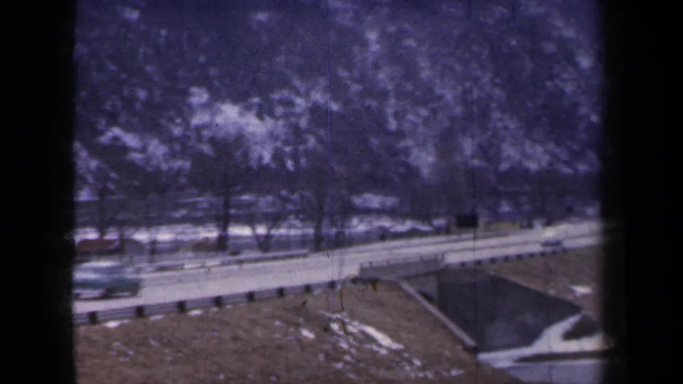 1959 New York 8mm frame with a car moving along a two-lane highway beside a bridge structure and snow-dusted mountain base.