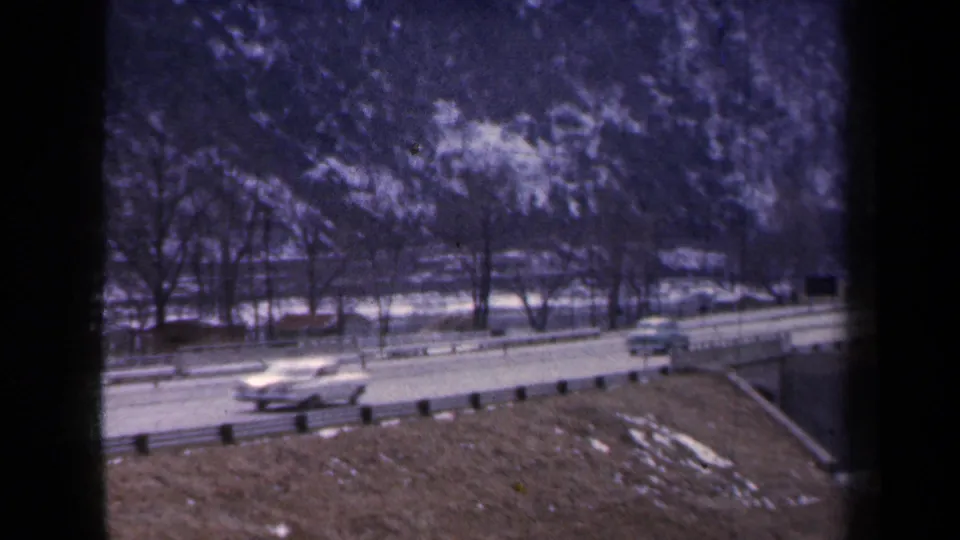 1959 New York 8mm frame showing mid-century cars on a two-lane highway beneath bare trees and a snowy mountain slope.