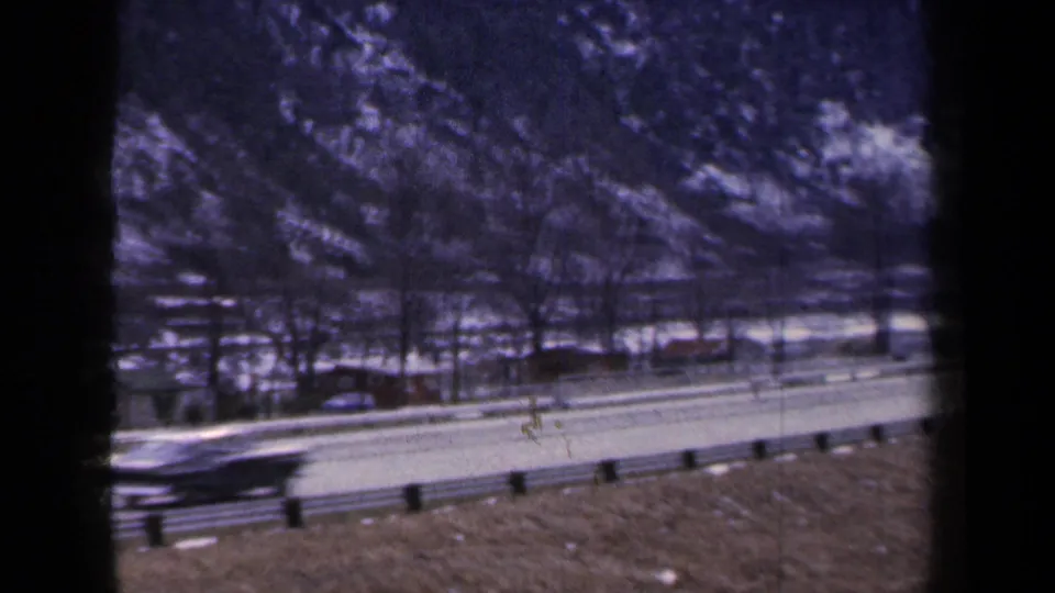 1959 New York 8mm frame with a motion-blurred vehicle passing on the highway below a snow-marked mountain base.