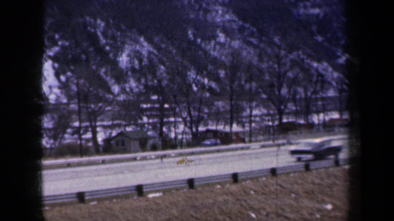 1959 New York home-movie frame showing a car on a snow-edged road with a guardrail and rocky hillside behind it.