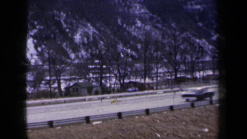 1959 New York home-movie frame showing a car on a snow-edged road with a guardrail and rocky hillside behind it.