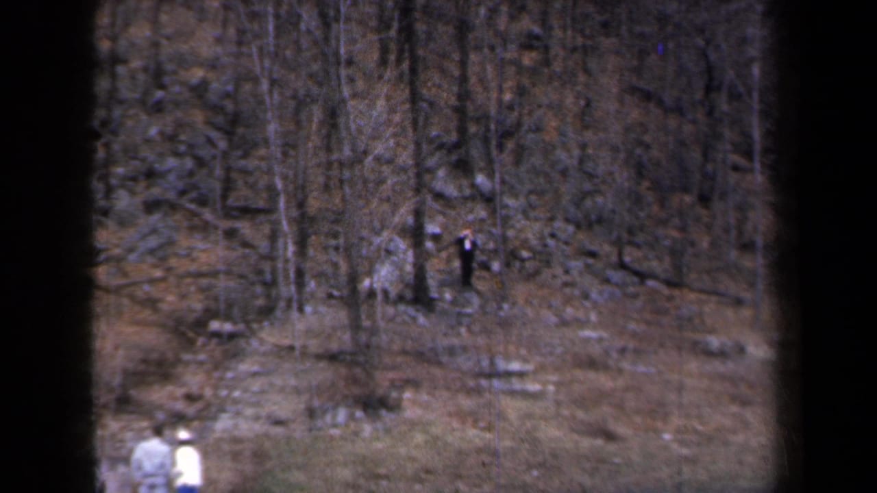1959 New York roadside view with bare trees, rocks, snow patches, and two distant pedestrians near the lower edge.