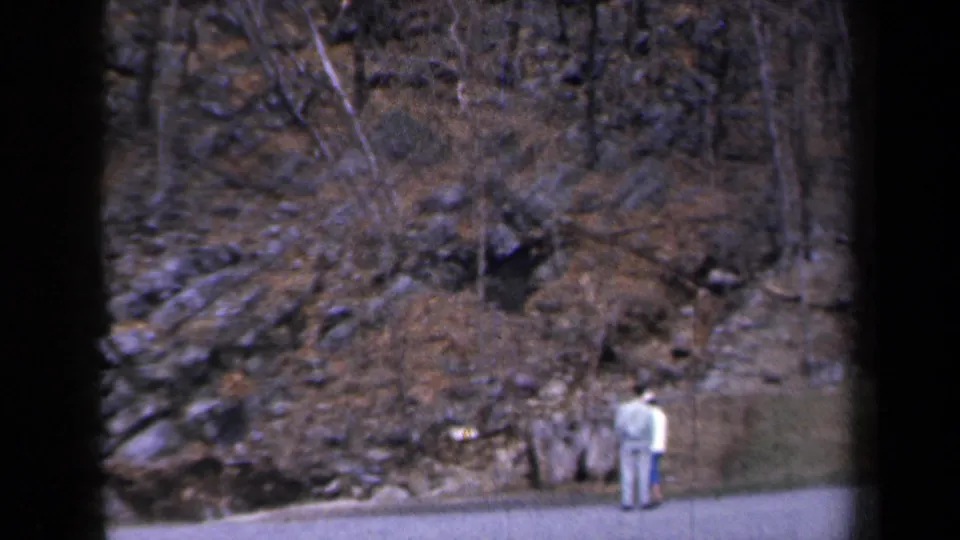 1959 New York home-movie frame showing two roadside figures below a rocky slope with bare trees and scattered snow.