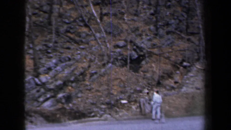 1959 New York roadside frame with two distant pedestrians, a paved edge, bare trees, rocks, and patchy snow on the hillside.