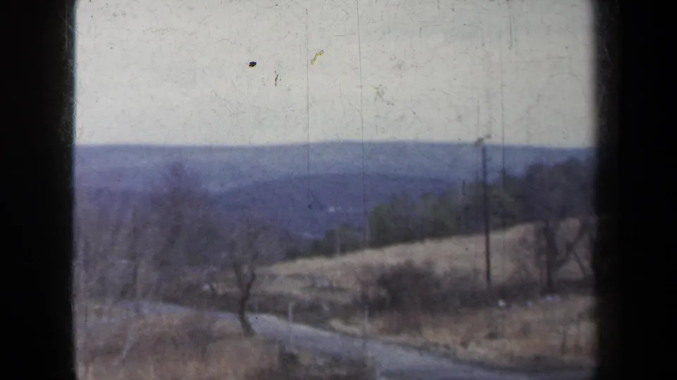 1959 New York 8mm frame with a rural road curving through bare trees, dry fields, utility poles, and distant hills.