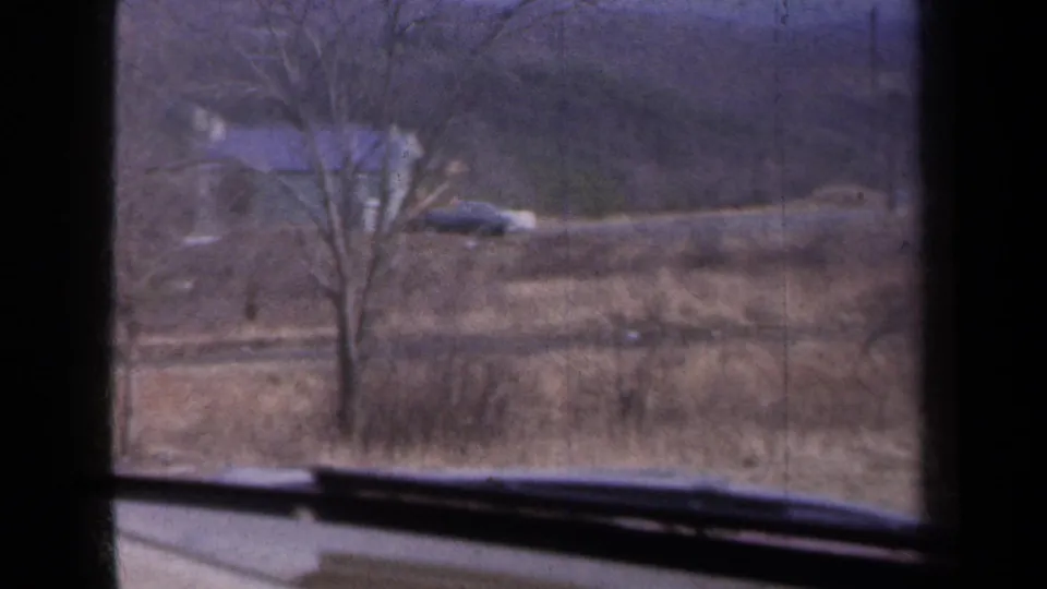 Rural New York countryside with a bare tree, brown hillside, and a distant light-roofed house seen through a vehicle window.