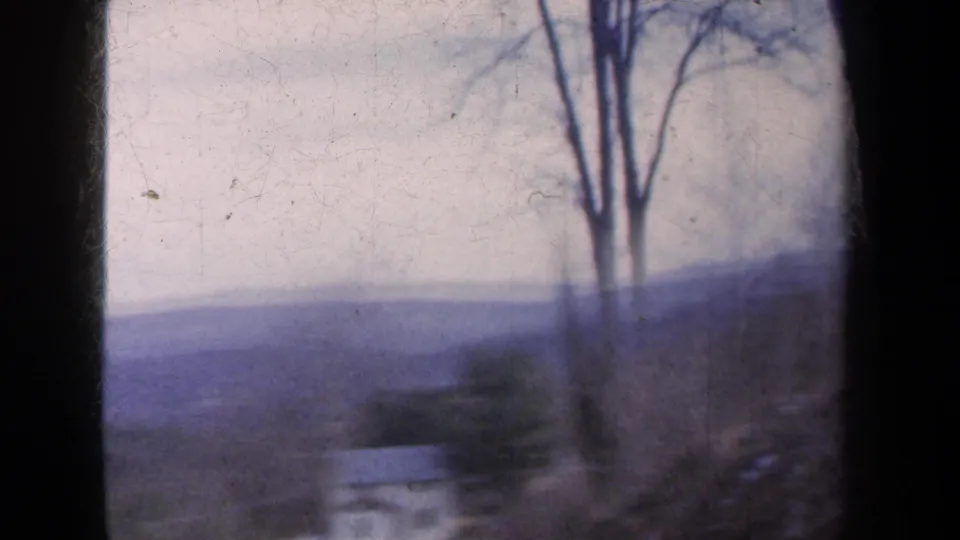 1959 New York 8mm frame showing bare roadside trees, distant purple hills, and dark window edges during a downhill vehicle view.