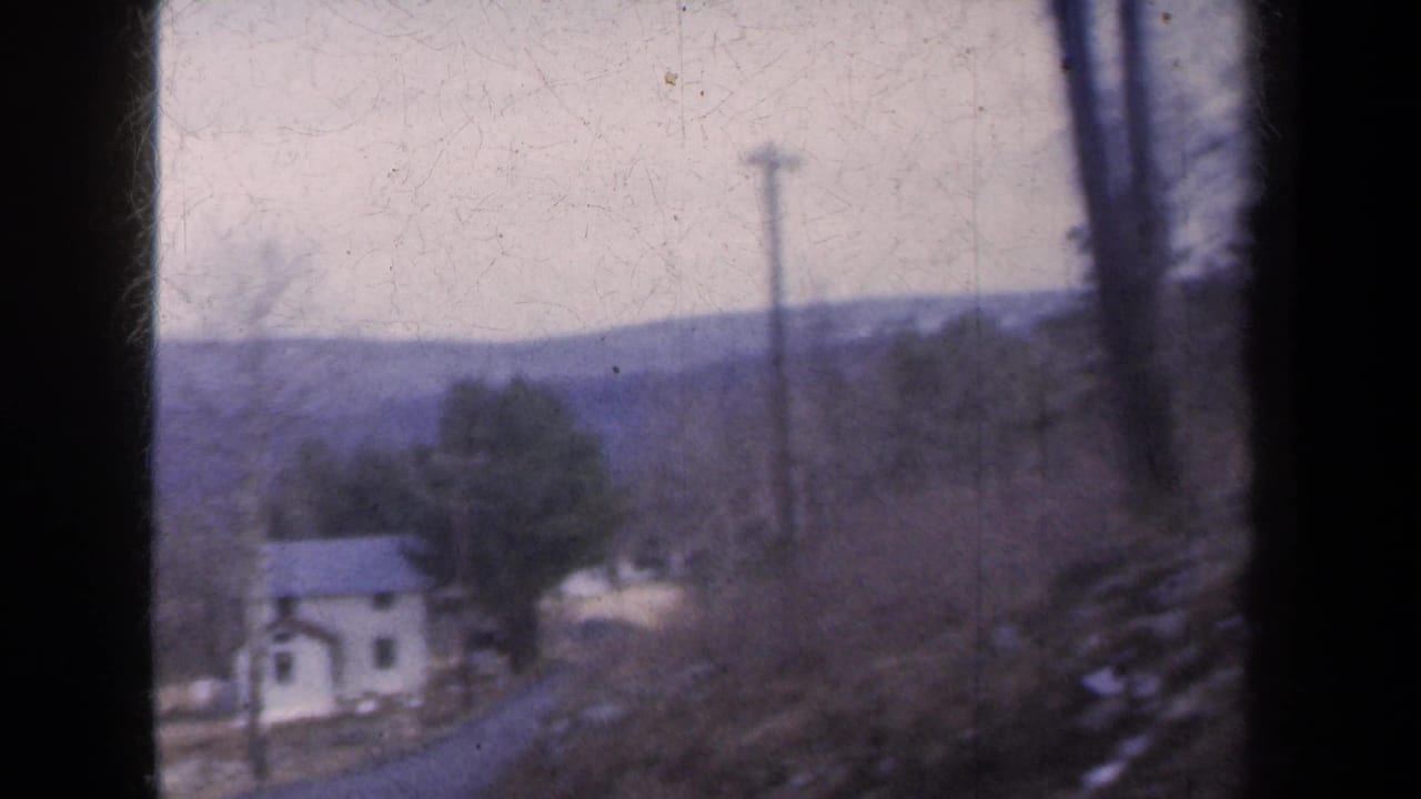 A white house with a dark roof appears among bare trees beneath distant hills in this 1959 New York home-movie frame.