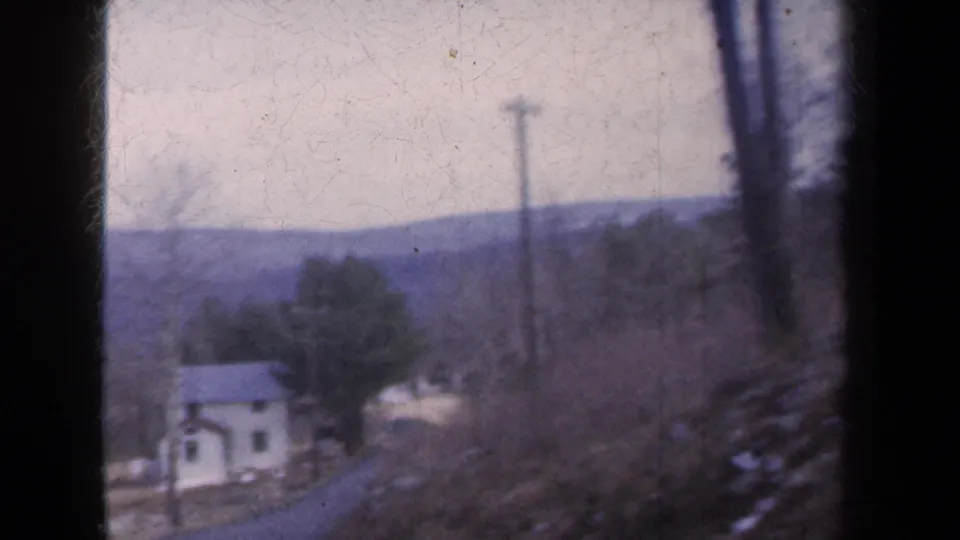 A white house with a dark roof appears among bare trees beneath distant hills in this 1959 New York home-movie frame.