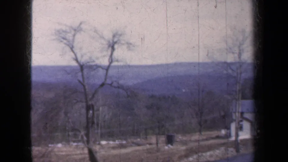 A jerky 8mm vehicle view shows roadside trees, utility poles, and a partial white building against distant New York hills.